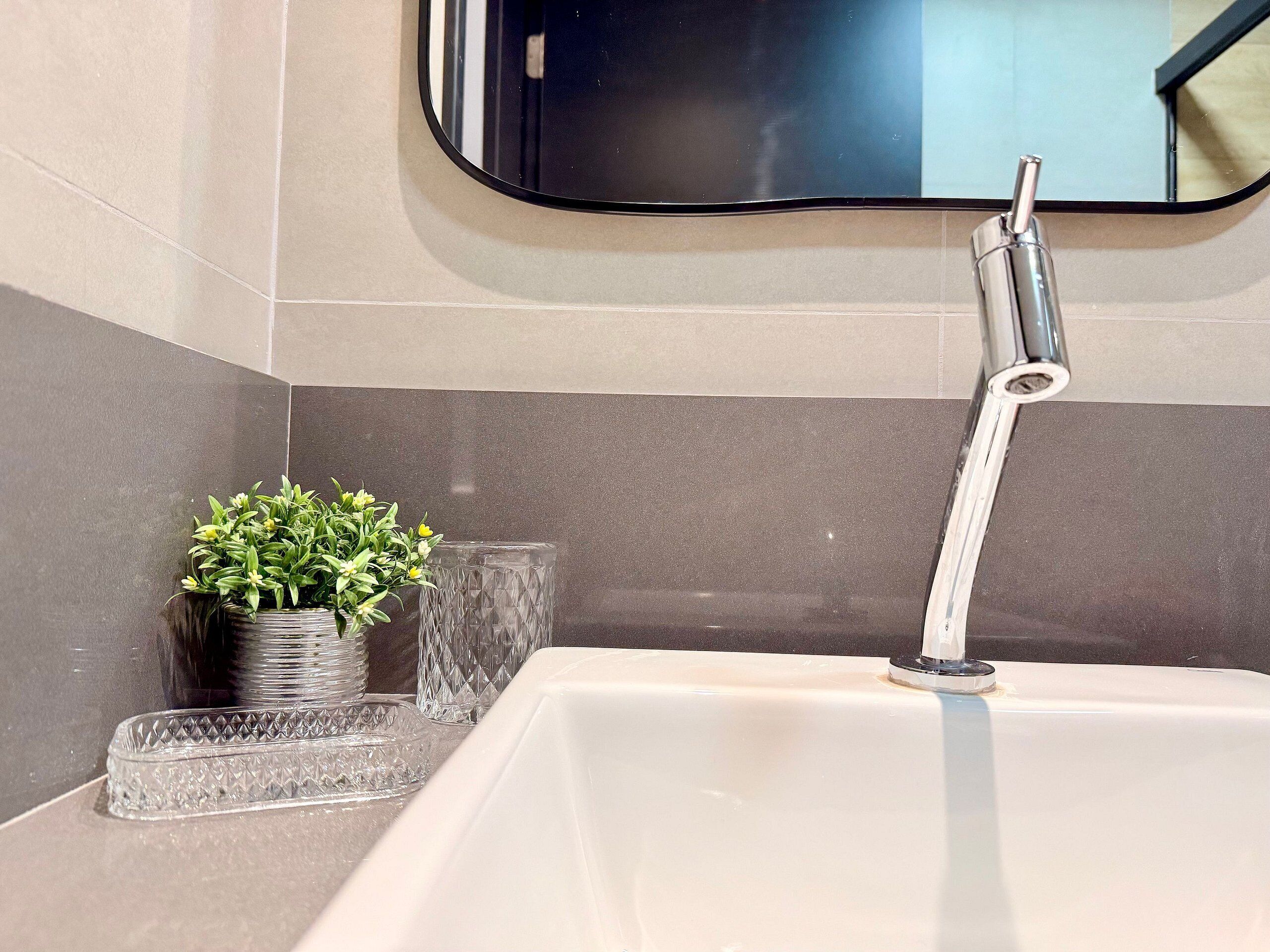 A bathroom with a white sink, a modern faucet, a small potted plant, and a glass container.