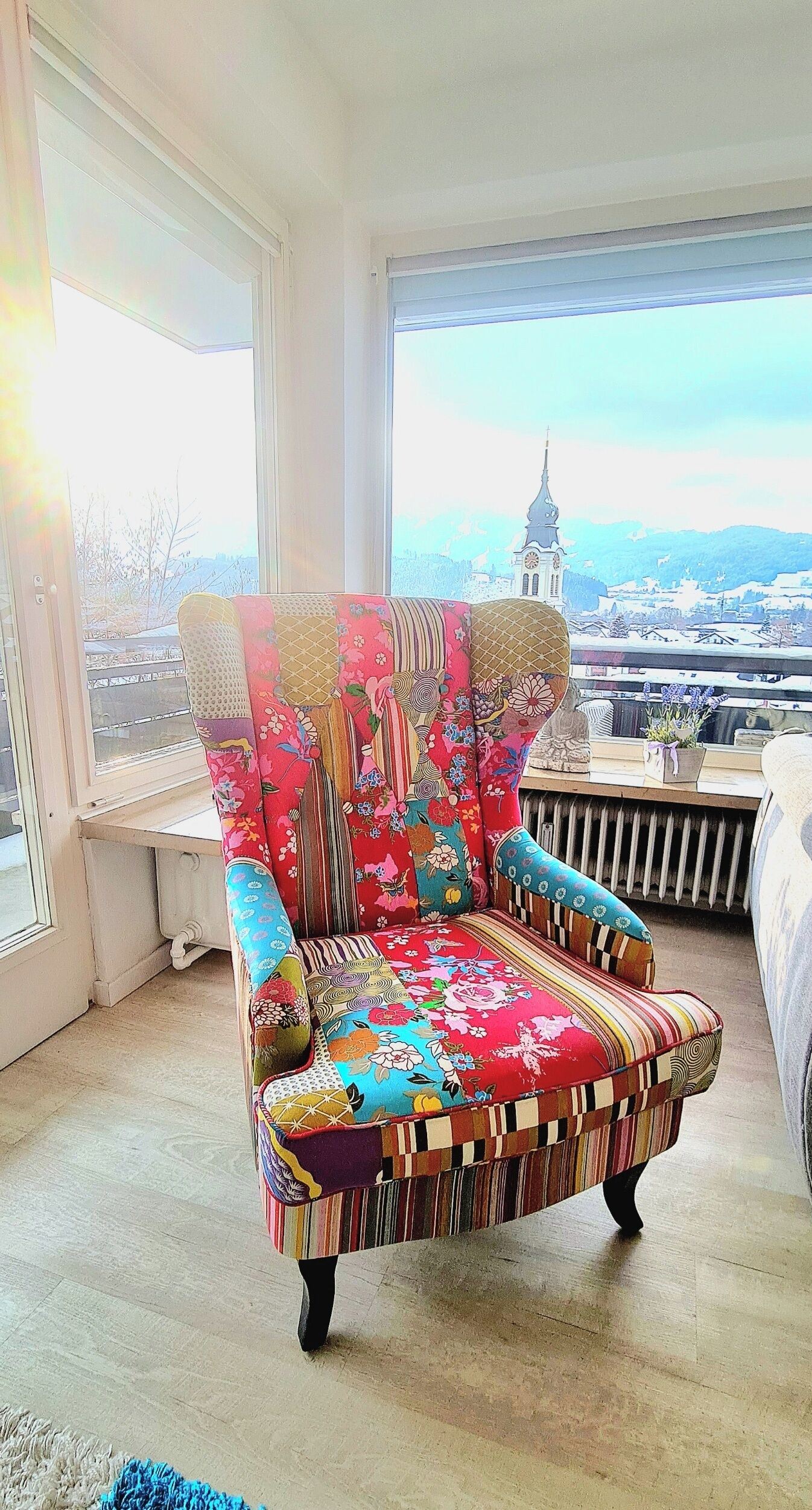 A colorful armchair with a floral pattern, positioned in a room with large windows overlooking a cityscape.