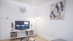 A modern living room with a flat-screen TV on a white console, a floor lamp, and abstract art on the wall. - 1-min walk to Town & Station I Near Airport I Parking (Luton)