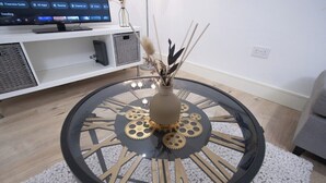 A round table with a decorative vase in the center, a television in the background, and a white shelf with storage boxes. - 1-min walk to Town & Station I Near Airport I Parking (Luton)
