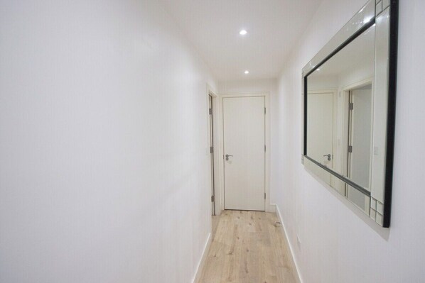 A narrow hallway with white walls and a wooden floor, leading to a door with a mirror on the right wall.