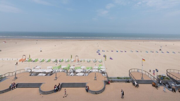 Een strand met een heldere lucht, een zandstrand en enkele mensen die wandelen. Er staan parasols en een vlaggenmast met een vlag.