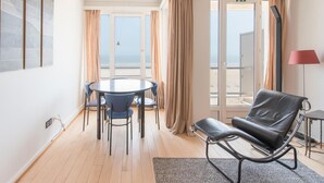 Een kamer met een ronde tafel, stoelen, een lederen bank, een lamp en uitzicht op zee door grote ramen.