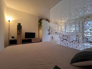 Ein Schlafzimmer mit einem großen Bett, einem Fernseher auf einem Holzbalken, einer dekorativen weißen Trennwand und einer Pflanze an der Wand.