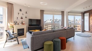 Ein modernes Wohnzimmer mit einer großen Couch, einem Kamin, einem Fernseher und Blick auf die Stadt durch bodentiefe Fenster.
