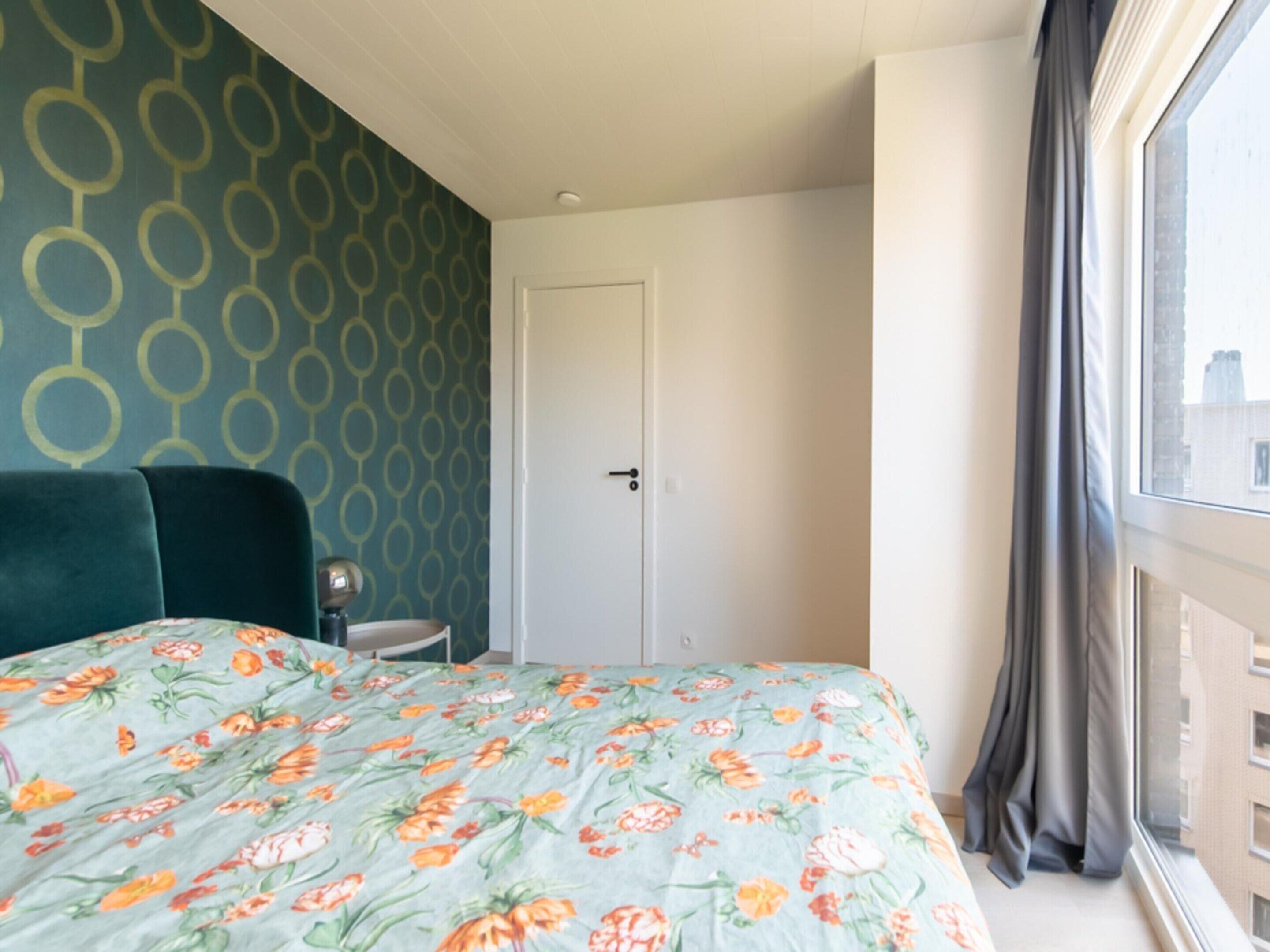 A hotel room with a floral bedspread, a green upholstered headboard, a white door, and a view of buildings through a window with curtains.