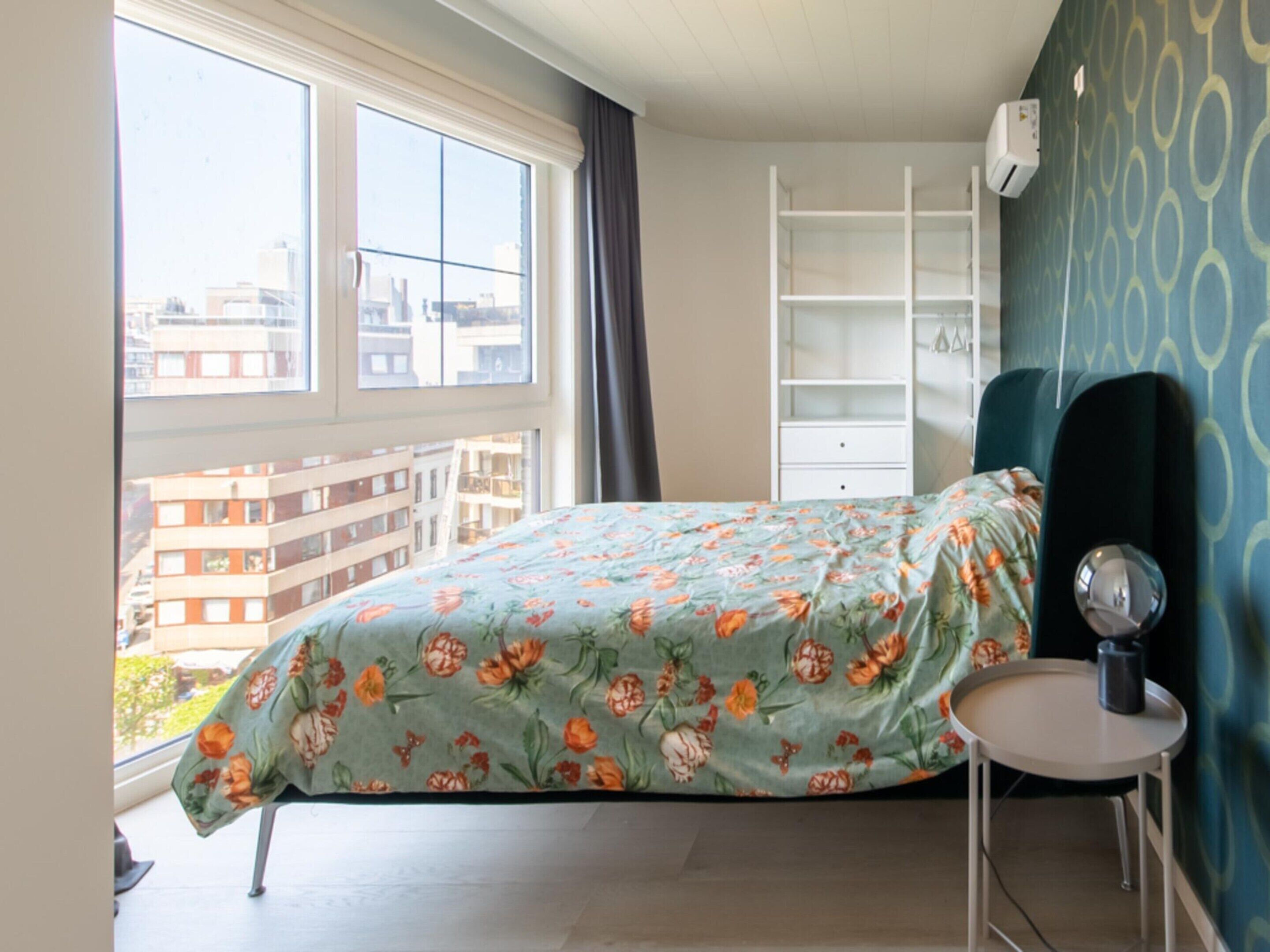 A bedroom with a bed, a small table, a wardrobe, and a window with a view of buildings.