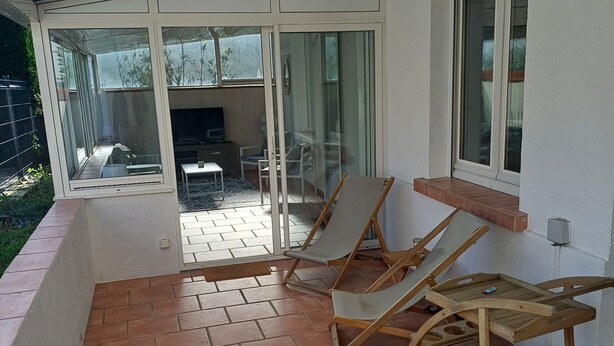 Eine überdachte Terrasse mit Fliesenboden, zwei Holzliegen, ein kleiner Holztisch und eine Glastür, die zu einem Außenbereich mit Tisch und Stühlen führt.