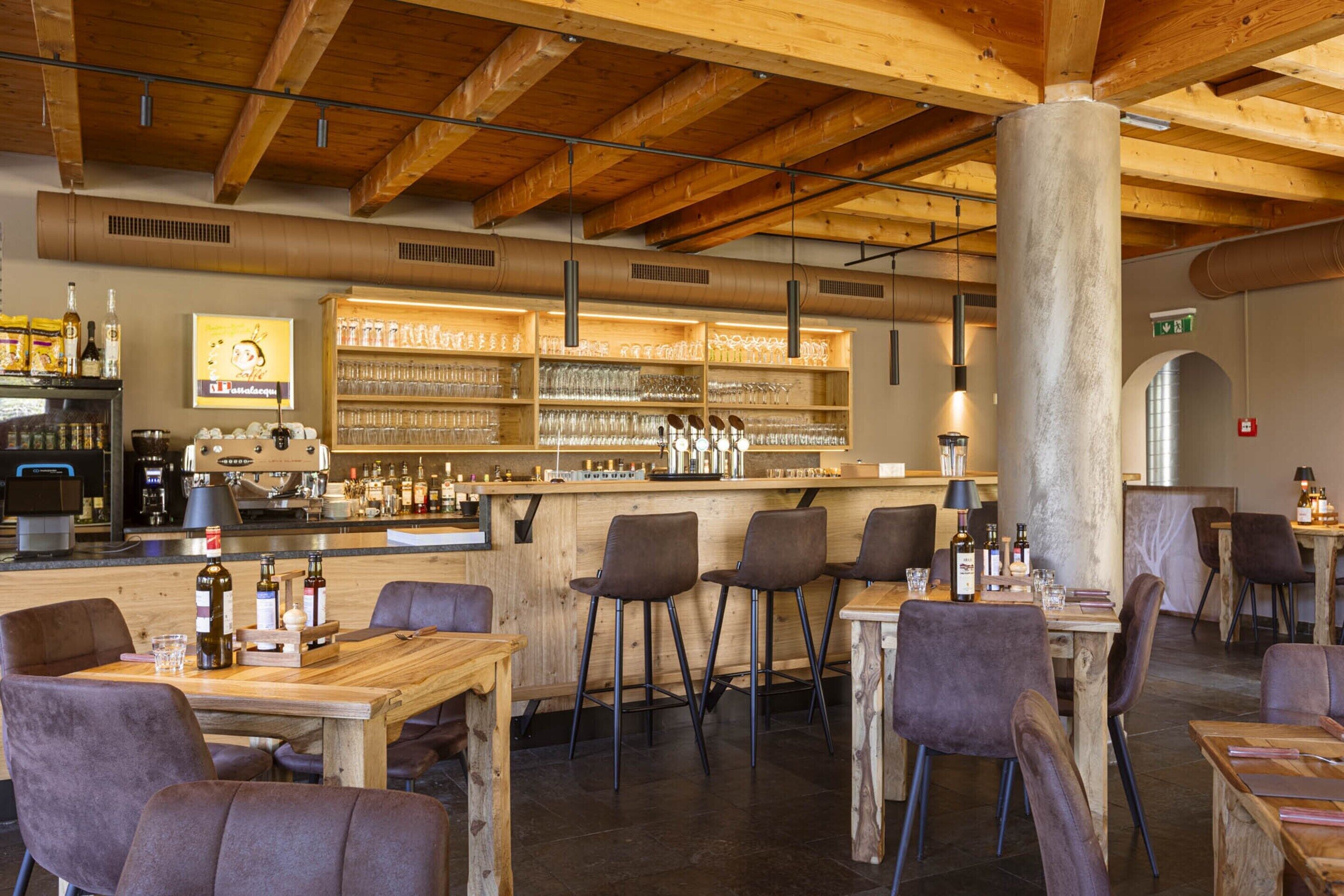 A restaurant with wooden tables and chairs, a bar area with shelves of glasses and bottles, and a high ceiling with exposed beams.