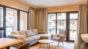 Un salon moderne avec un canapé d’angle, une table basse ronde et deux fauteuils. De grandes fenêtres offrent une vue sur l’extérieur du bâtiment.