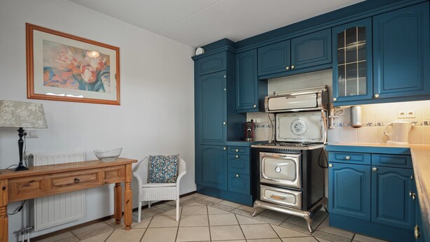 Een keuken met blauwgroene kastjes, een houten tafel, een witte stoel en een retro fornuis.