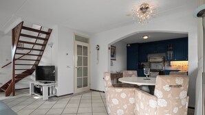 Een woonkamer met een televisie, fauteuils met bloemenprint, een trap en een keuken met blauwe kasten.