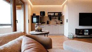A modern living room with a sofa, a coffee table with a decorative item, a kitchen area with cabinets and appliances, and a TV mounted on the wall. - SeeZeit at Walchsee (Walchsee)