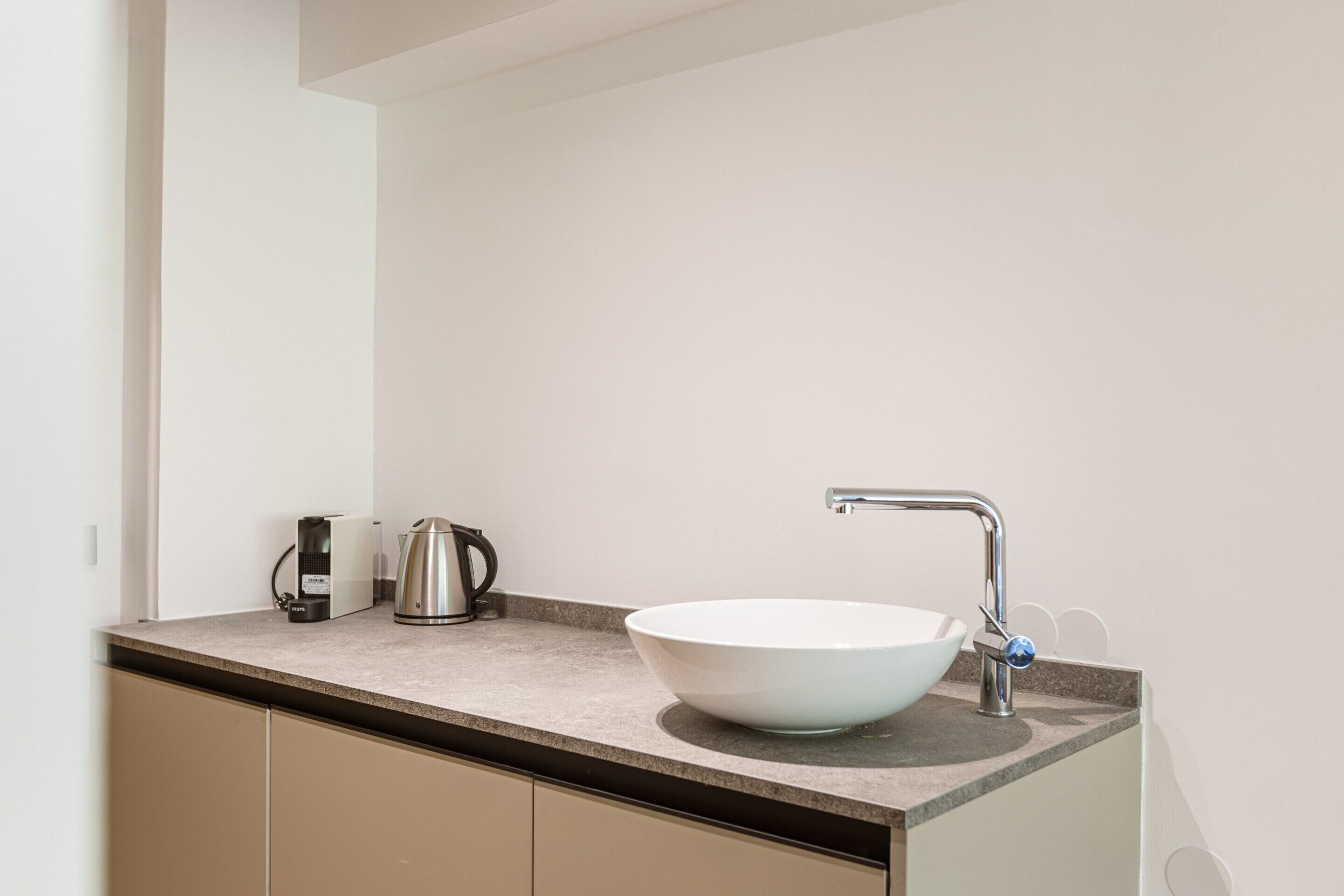 A modern bathroom with a white sink, a stainless steel kettle, and a coffee maker on the countertop.