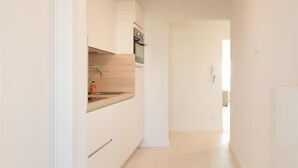 Um corredor estreito e bem iluminado, com paredes brancas e chão de azulejos, que dá acesso a uma cozinha com armários brancos e bancada de madeira.