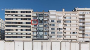 Um hotel com vários andares e varandas, uma praia ao primeiro plano e um céu claro.