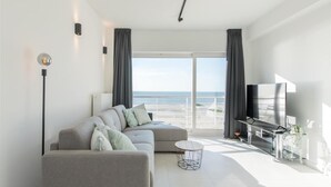 Uma sala de estar moderna com um sofá cinzento, uma mesa de centro redonda, uma televisão de tela plana e uma grande janela com vista para a praia.