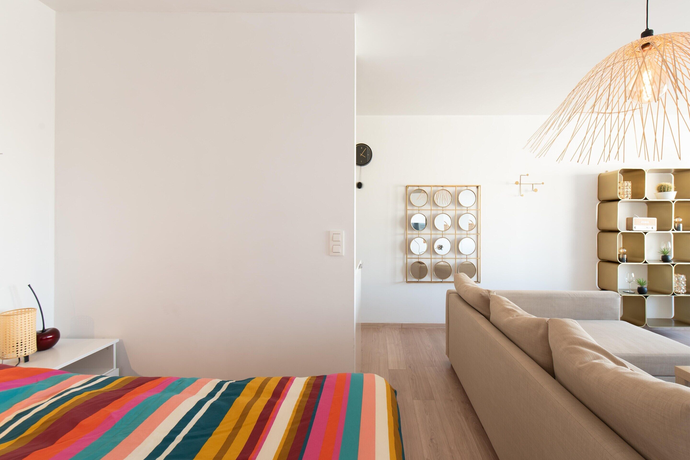 Ett modernt hotellrum med en randig säng, en beige soffa, en väggklocka och en dekorativ väggpanel.