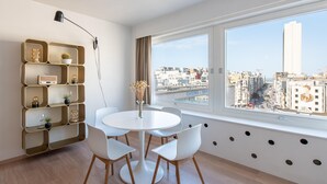 Una habitación moderna con una mesa redonda, sillas blancas, una estantería con marco dorado y un ventanal con vistas a la ciudad.