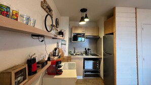 Cocina compacta con microondas, horno y refrigerador. Se observan estantes con diversos utensilios de cocina y una cafetera.