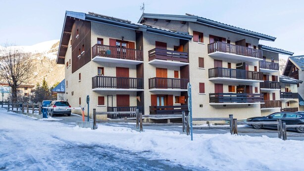 A multi-story building with balconies, a ski slope in the background, and a snow-covered ground.