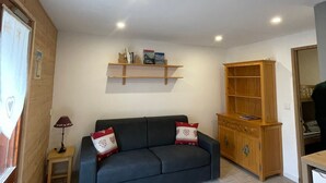 A living room with a dark blue sofa, two decorative pillows, a small wooden side table with a lamp, a wooden cabinet, and a wall-mounted shelf with books and decorative items.