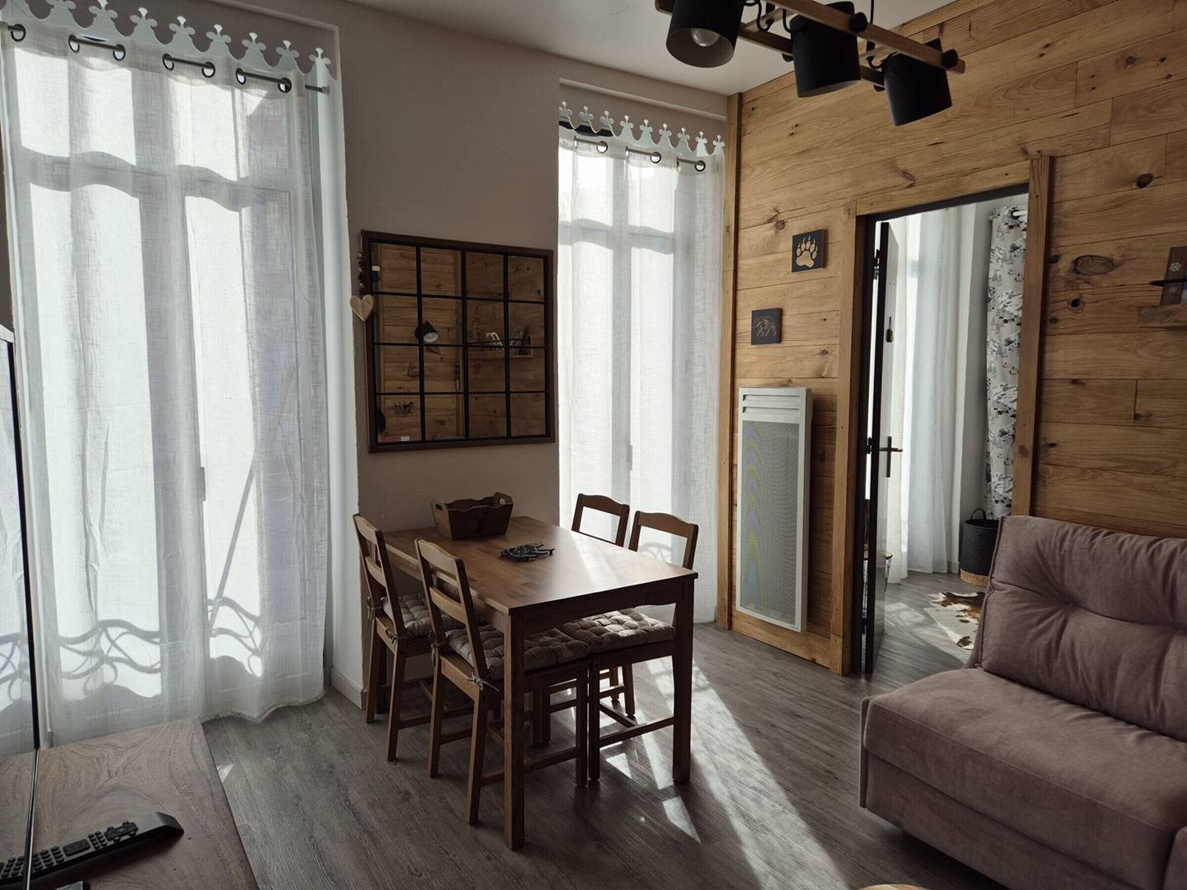Una sala de estar acogedora con piso de madera, un sofá marrón, una mesa de comedor con sillas y un ventanal con cortinas translúcidas.