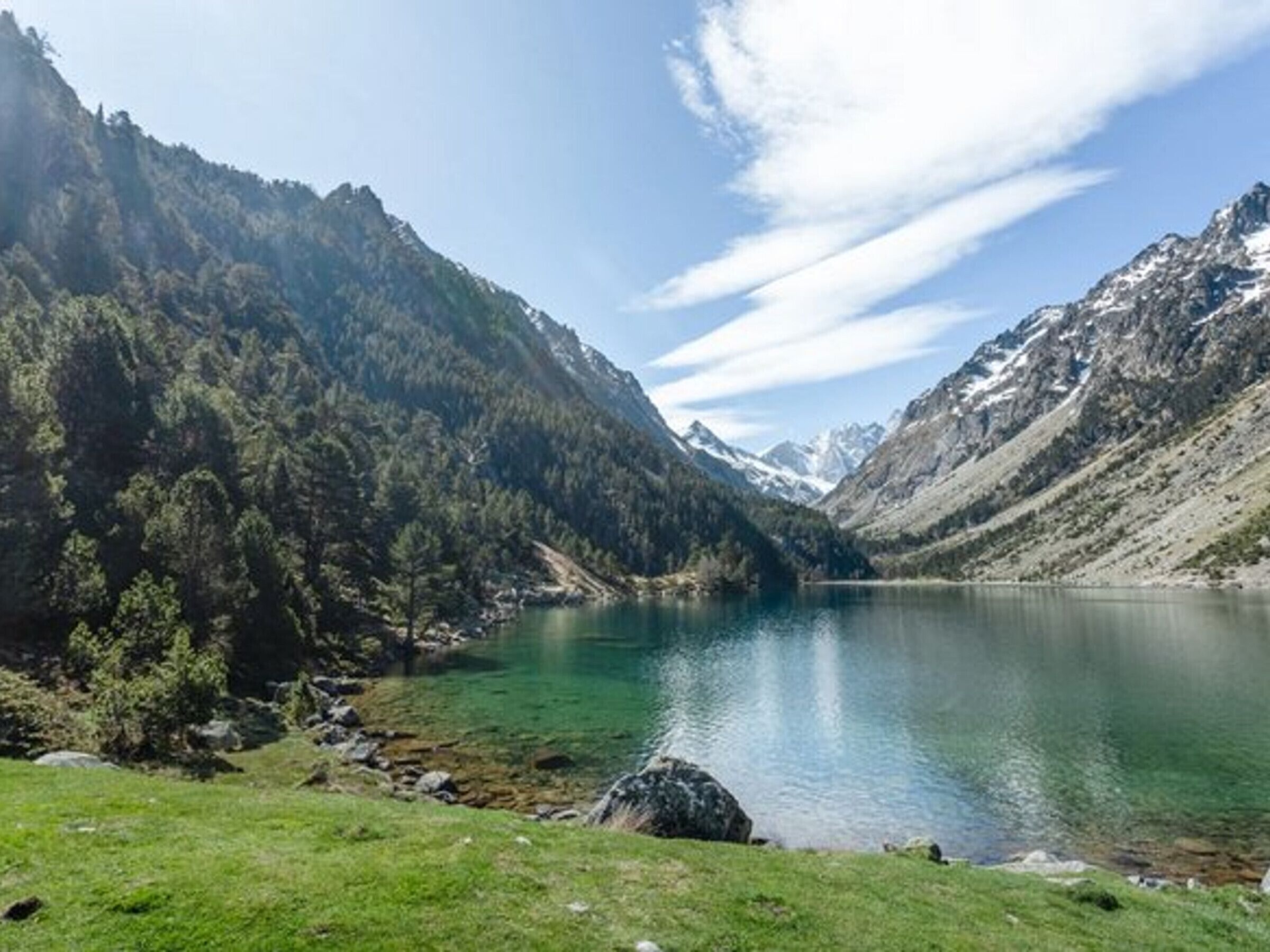 Ein Bergsee mit klarem Wasser, umgeben von bewaldeten Hügeln und blauem Himmel.