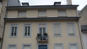 A two-story building with a blue door, multiple windows with shutters, and a balcony with potted plants.