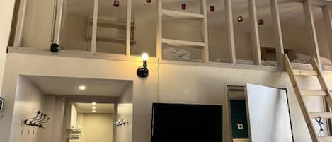 Une pièce avec un téléviseur fixé au mur, une échelle et un espace en mezzanine en cours de construction, avec des poutres apparentes et des lumières de sécurité rouges.