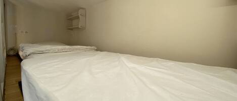 Une chambre avec un lit simple, un plancher en bois, un mur blanc et une petite étagère blanche.