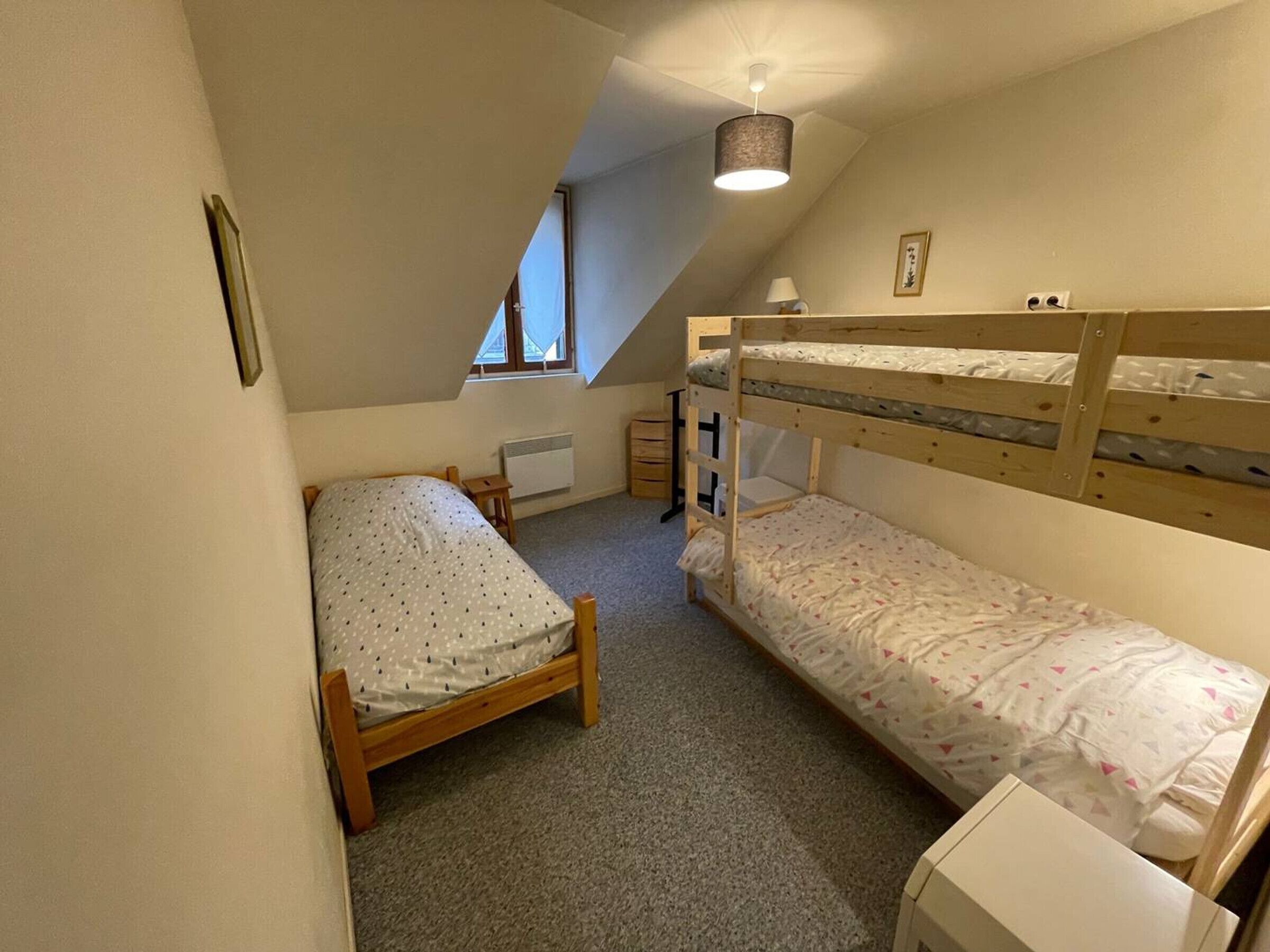 Una habitación pequeña y acogedora con techo inclinado, una cama litera, una cama individual, un radiador, una mesita y una lámpara en el techo.