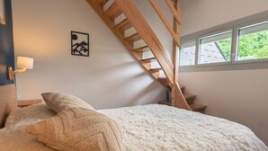 A bedroom with a bed, pillows, a nightstand, a lamp, a wooden staircase, and a window with a view of greenery.