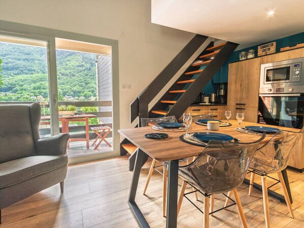 Une cuisine moderne dotée d’un four encastré, un coin repas avec une table et des chaises en bois, et une vue sur un paysage boisé grâce à de grandes fenêtres.