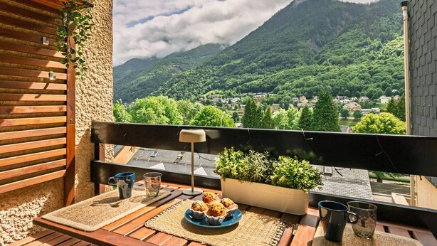 A balcony with a view of a mountain, a table with a plate of muffins, and a potted plant.