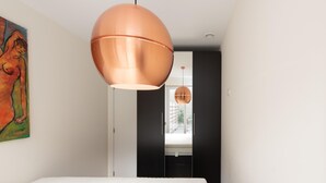 Ein modernes Schlafzimmer mit einem großen Bett, einer hängenden Kupferdpendelleuchte und Blick auf eine Stadtkulisse durch ein Fenster.