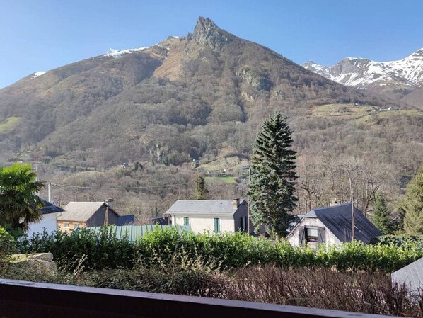 Una montaña con un pico destacado, rodeada de casas con techos inclinados, y un alto pino en primer plano.