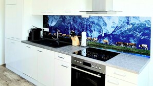 Eine moderne Küche mit weißen Schränken, einem Edelstahlherd und einer großen Kochinsel mit Wandverkleidung im Bergmotiv.