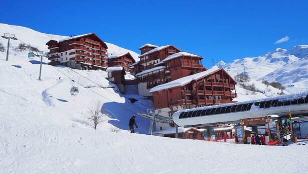 A snow-covered mountain resort with wooden buildings, ski lift, and a clear blue sky.