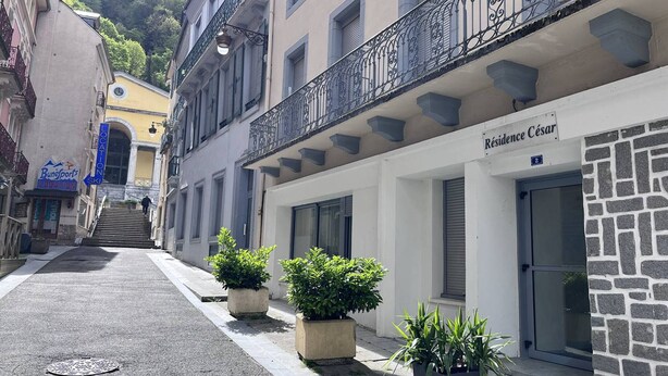 A narrow street with buildings on either side, including a hotel named 'Résidence César' with a stone facade and a balcony with wrought iron railings.