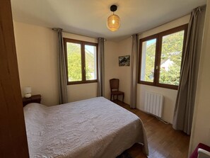A bedroom with a bed, two windows, a chair, a radiator, and a hanging light fixture. - Spacious House in Cauterets: 4 Bedrooms, Garage, No Pets (Cauterets)