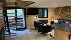 A modern kitchen with a wooden staircase, a dining area with a black table and chairs, and a living area with a sofa and a fireplace.
