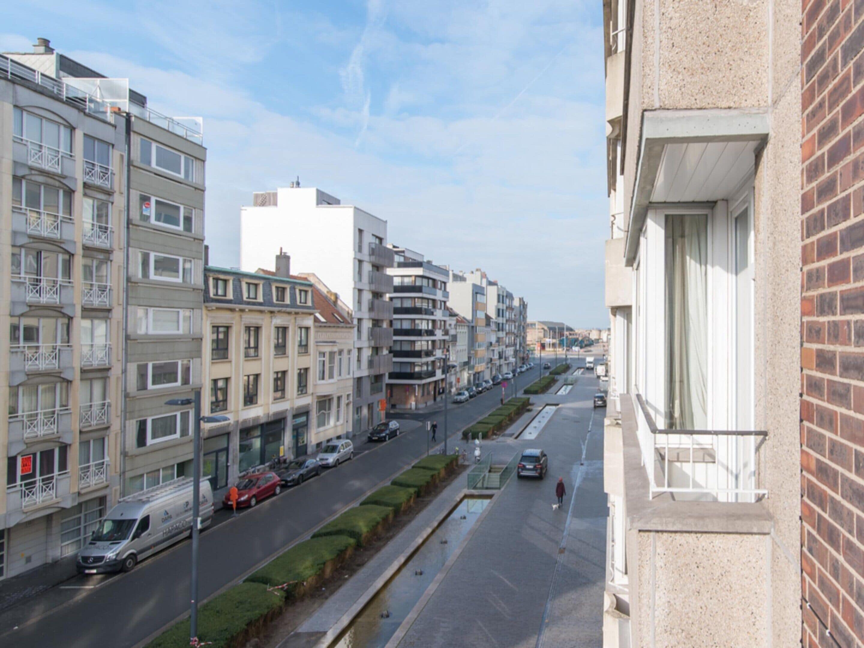 A multi-story residential building with balconies, a narrow street with parked cars, and a pedestrian pathway.