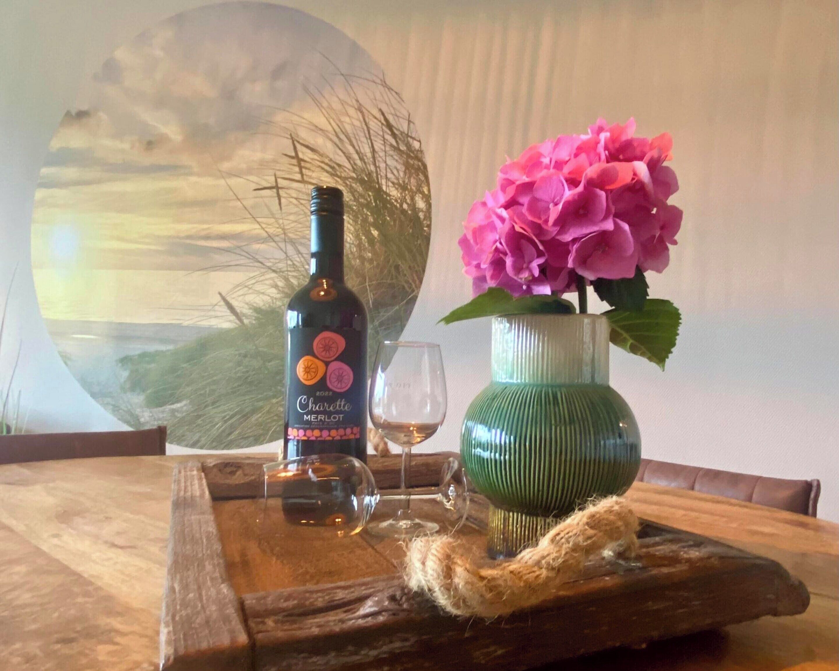 A bottle of 2012 Charette Merlot wine, a glass of wine, and a vase with pink flowers on a wooden tray.