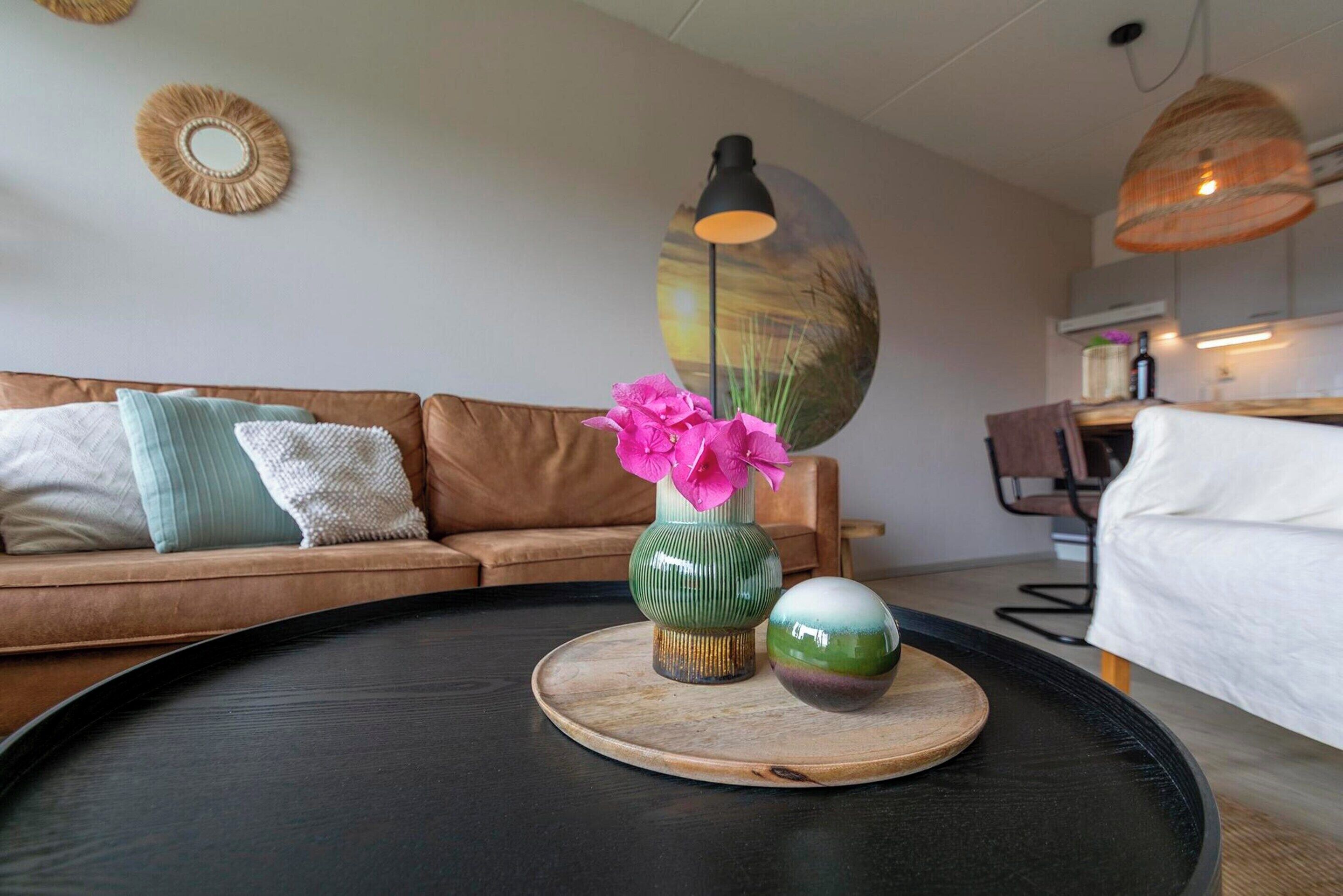 A modern living room with a brown sofa, a round coffee table, a vase with pink flowers, and a wall-mounted lamp.