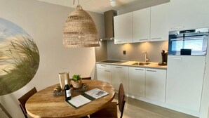 A modern kitchen with white cabinets, a built-in oven, and a dining area with a round wooden table and leather chairs. - Idyllic Coastal Base w/ Garden (Callantsoog)