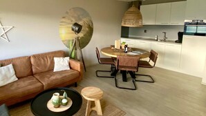 A modern living room with a brown leather sofa, a round dining table with chairs, a small wooden stool, and a black coffee table with decorative items. - Idyllic Coastal Base w/ Garden (Callantsoog)