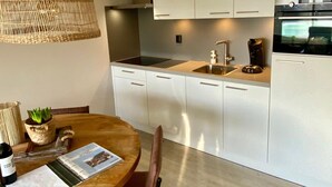 A modern kitchen with white cabinets, a stainless steel sink, and an oven. There is a wicker pendant light above the dining table, which has a magazine and a bottle on it. - Idyllic Coastal Base w/ Garden (Callantsoog)