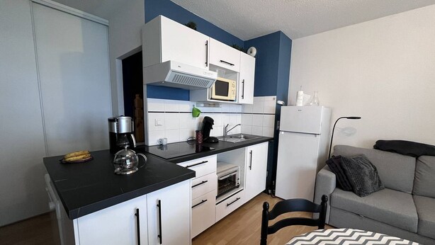 A compact kitchen with white cabinets, a microwave, a coffee maker, and a toaster. There's a grey sofa and a patterned rug in the living area. - Cozy apartment in Cauterets with terrace and ski storage (Cauterets)