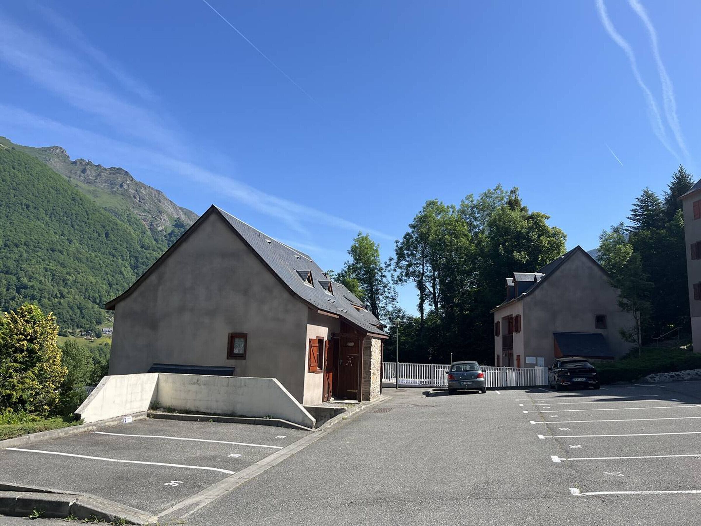 A parking lot with two buildings, one with a sloped roof and another with a flat roof, surrounded by greenery and mountains.
